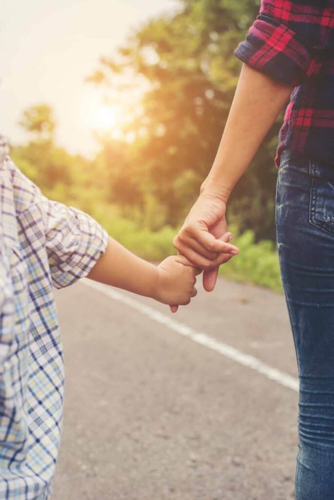 Mother holding hand of her son outdoors in summer walking on the roadside.