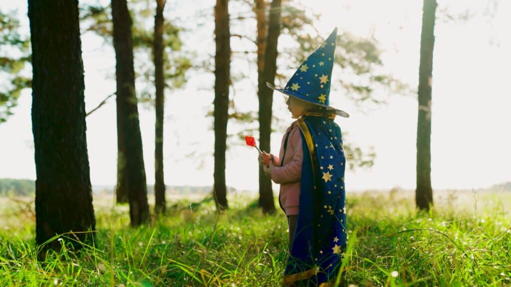 Girl, child plays in wizard costume, holds toy with a magic wand in park.