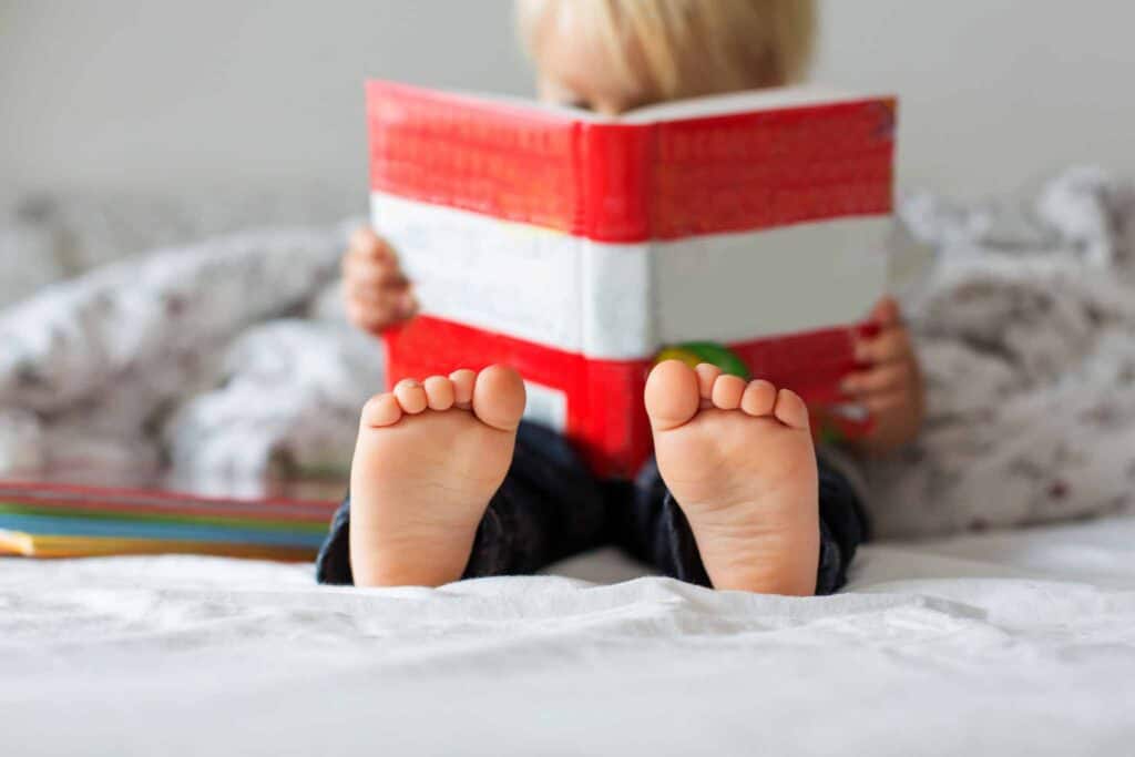 Sweet toddler boy, reading book at home, sitting in bed, lots of books around him